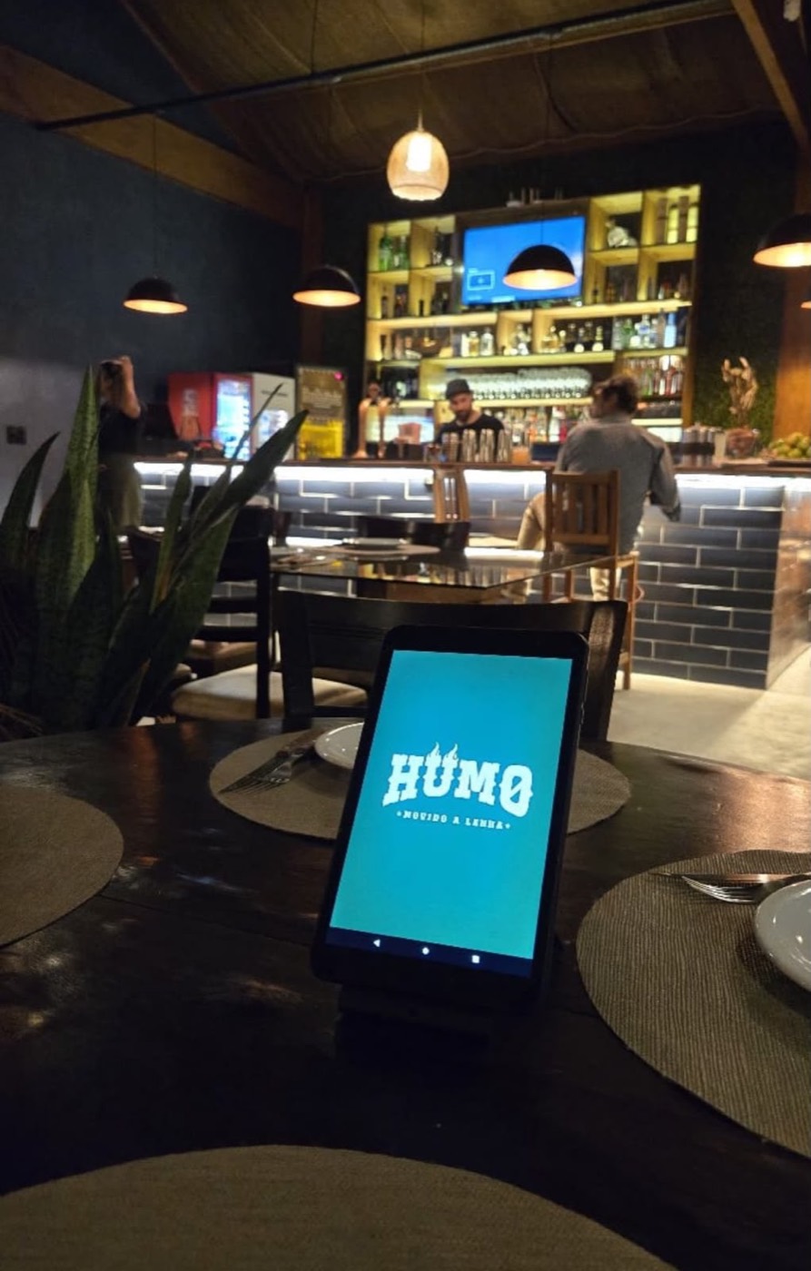 Tablet em pé na mesa de um restaurante à noite. A tela azul mostra a marca Humo. Ao fundo, o bar com prateleira de garrafas e luminárias.